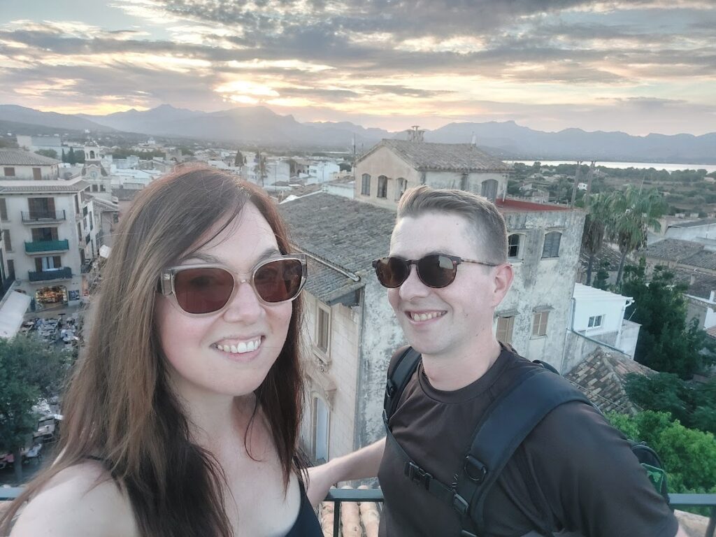 Two people wearing sunglasses smile on a rooftop with a scenic view of a sunset over a historic Mediterranean town.